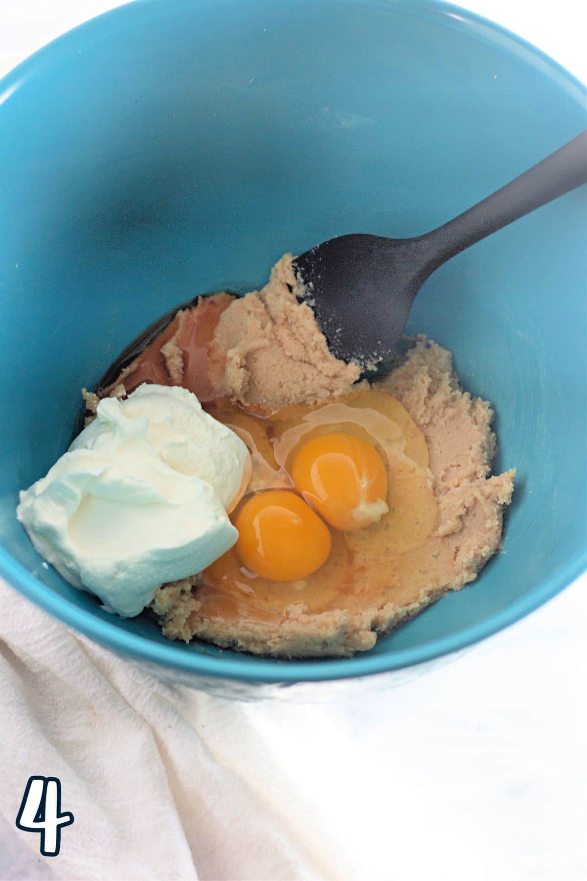 A blue bowl with sour cream, eggs, butter, and sugar with a spoon. 