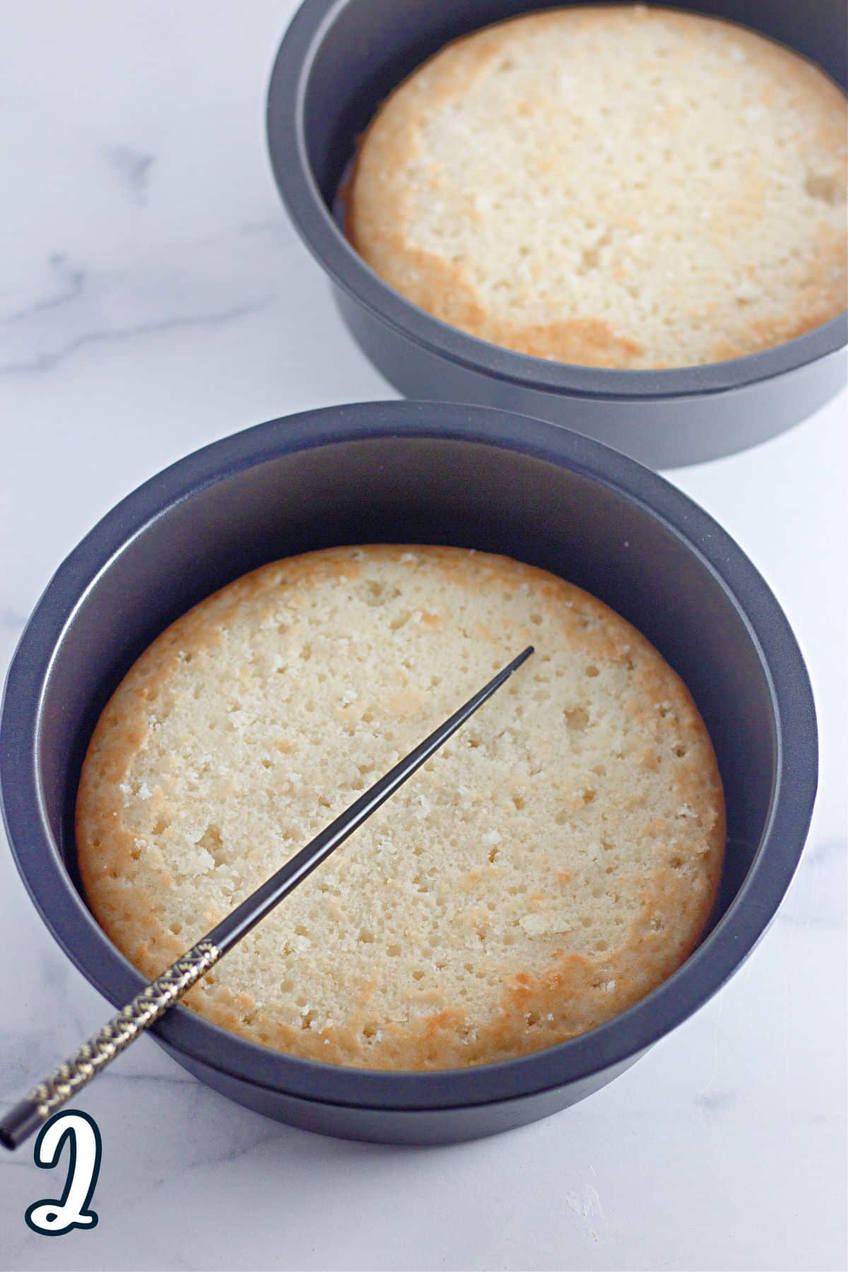 Two baked white cakes still in the pan with a chop stick used to poke holes in the cakes.