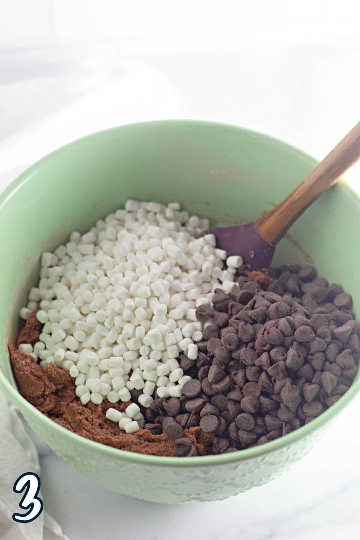 Chocolate chips, dried marshmallow, and cookie dough in a bowl with a wooden spoon.