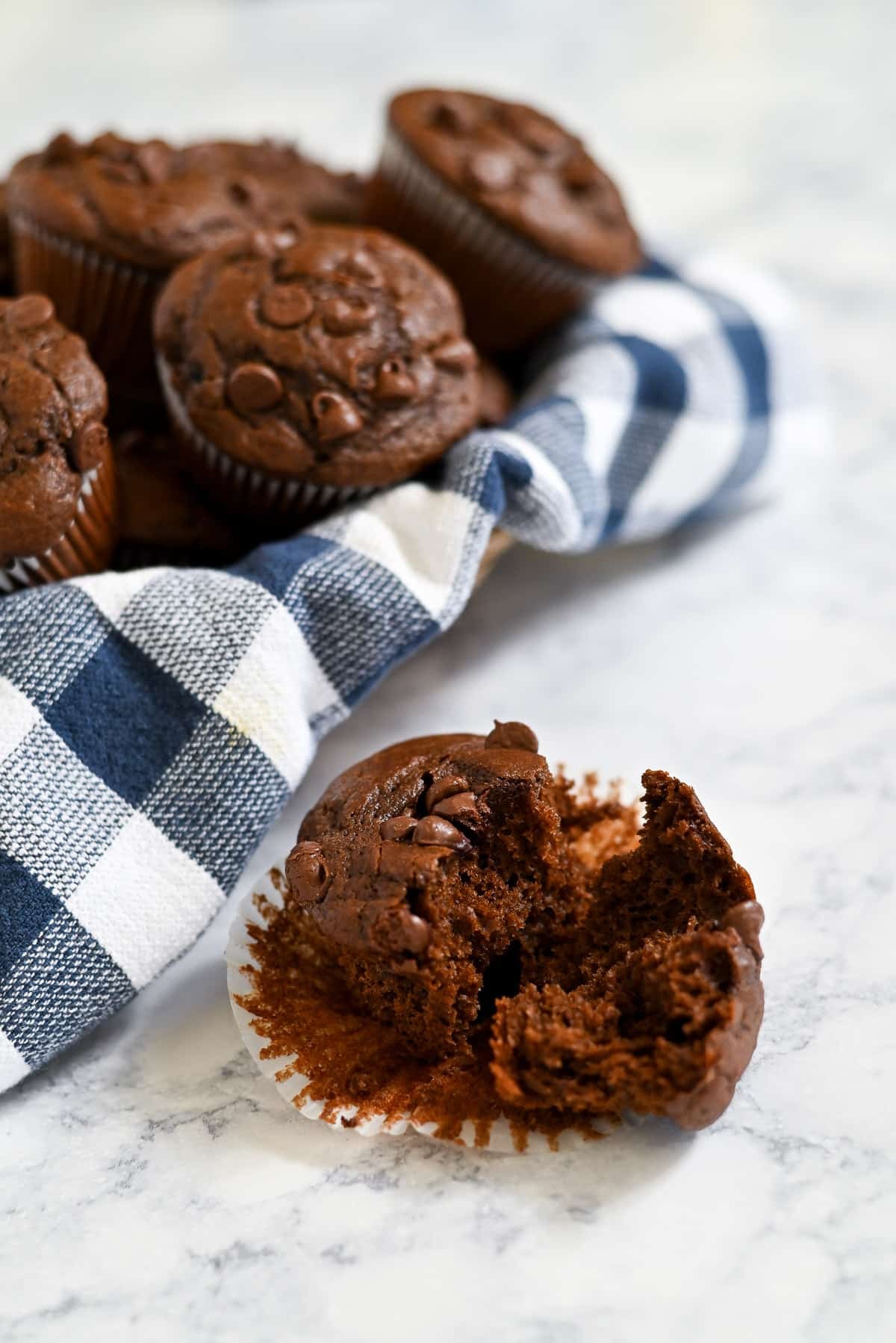Chocolate Espresso Muffins with chocolate chips rest on a blue plaid cloth; one muffin is half-eaten on a marble surface.