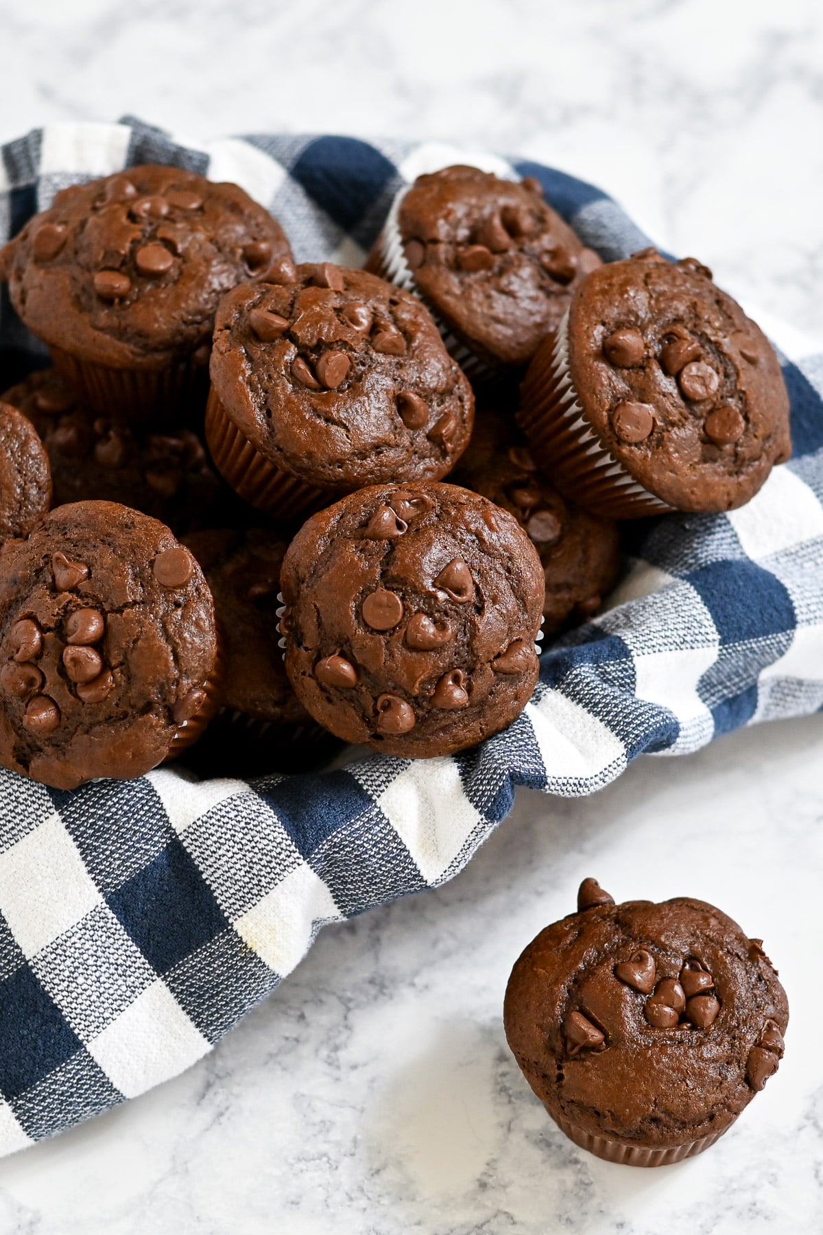 A pile of Chocolate Espresso Muffins on a blue and white checkered cloth, with some muffins scattered nearby.