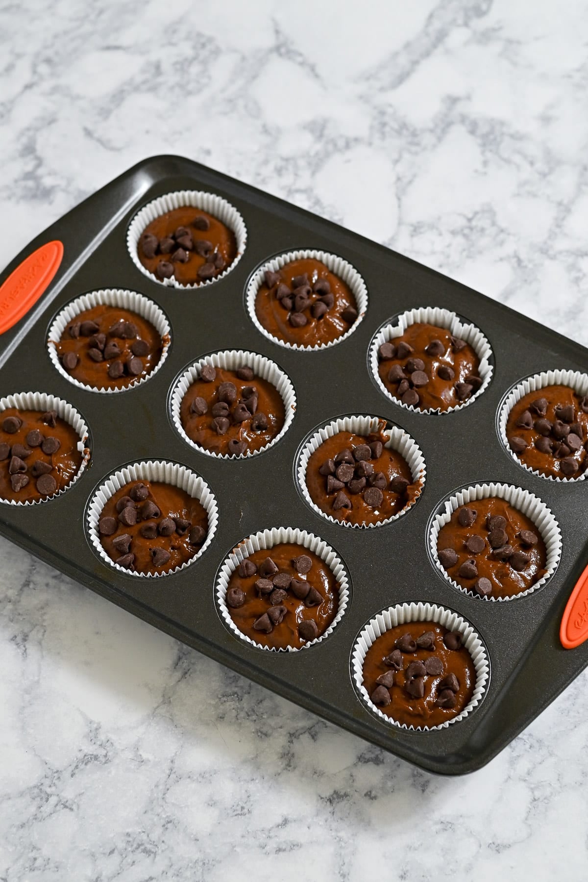 A muffin tin filled with rich Chocolate Espresso Muffins batter and chocolate chips, ready to be baked, sits on a marble countertop.