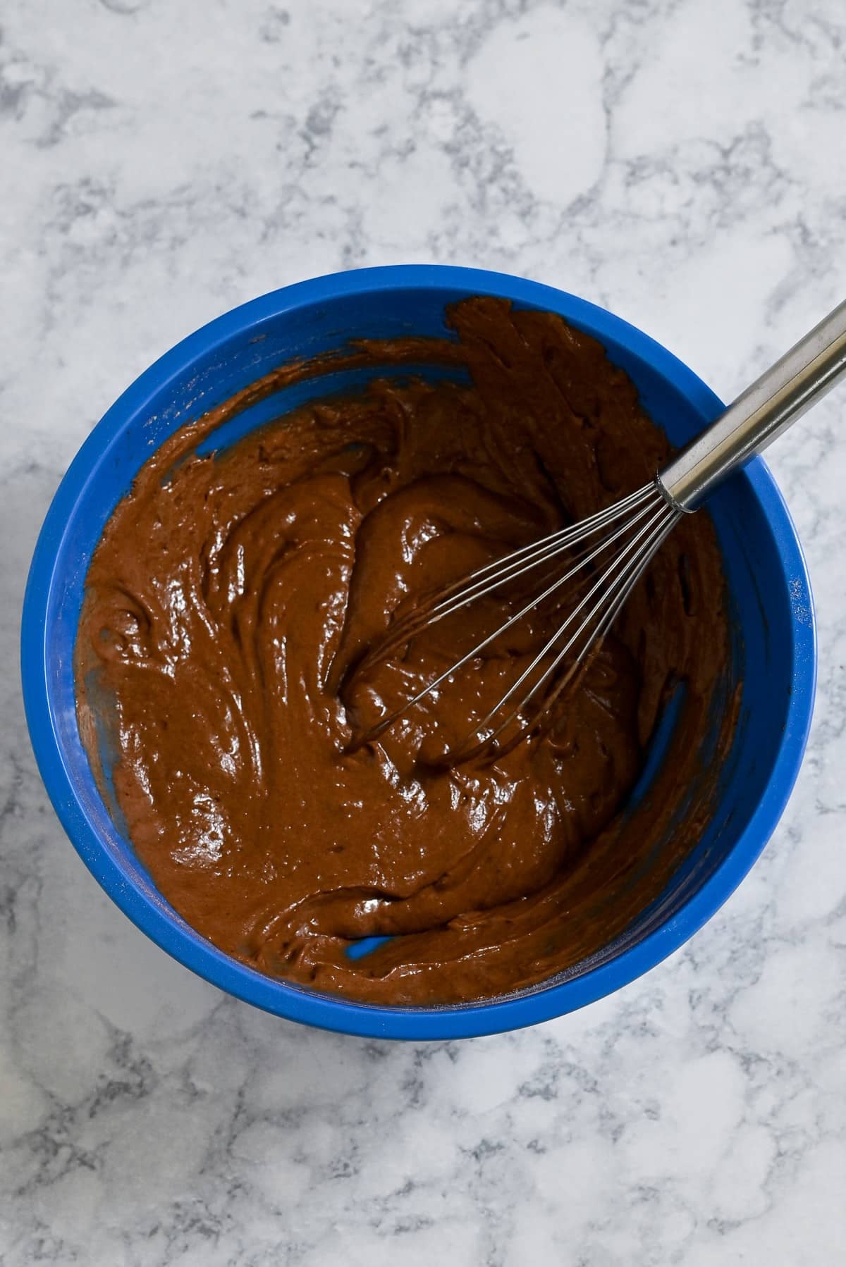 A blue bowl with chocolate cake batter for Chocolate Espresso Muffins and a whisk on a marble countertop.