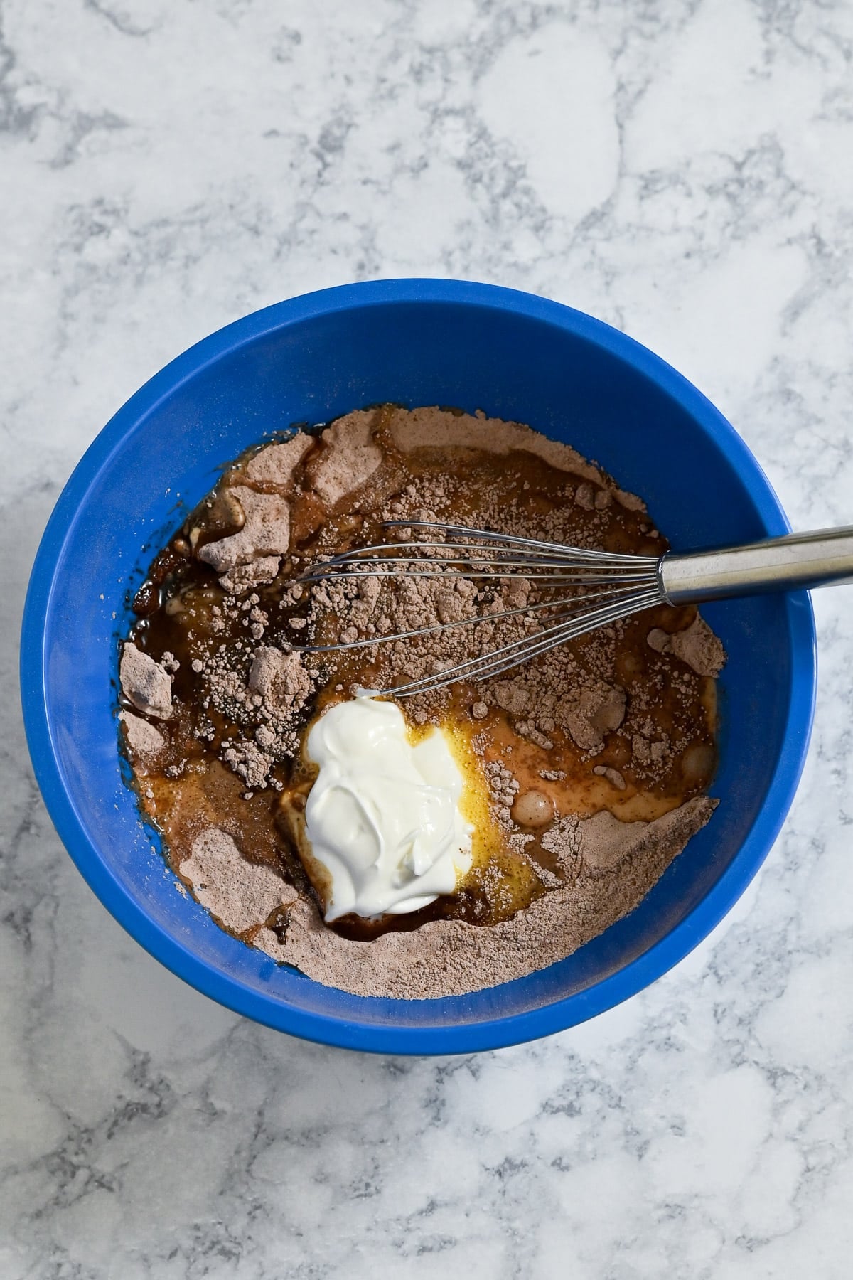 Blue bowl with cake mix, yogurt, eggs, and a whisk on a marble countertop, ready to be mixed for delicious Chocolate Espresso Muffins.