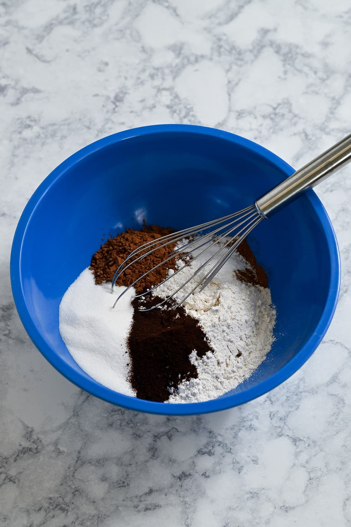 A blue bowl with sugar, flour, cocoa powder, coffee, and a whisk on a marble countertop-everything you need to start baking delicious Chocolate Espresso Muffins.