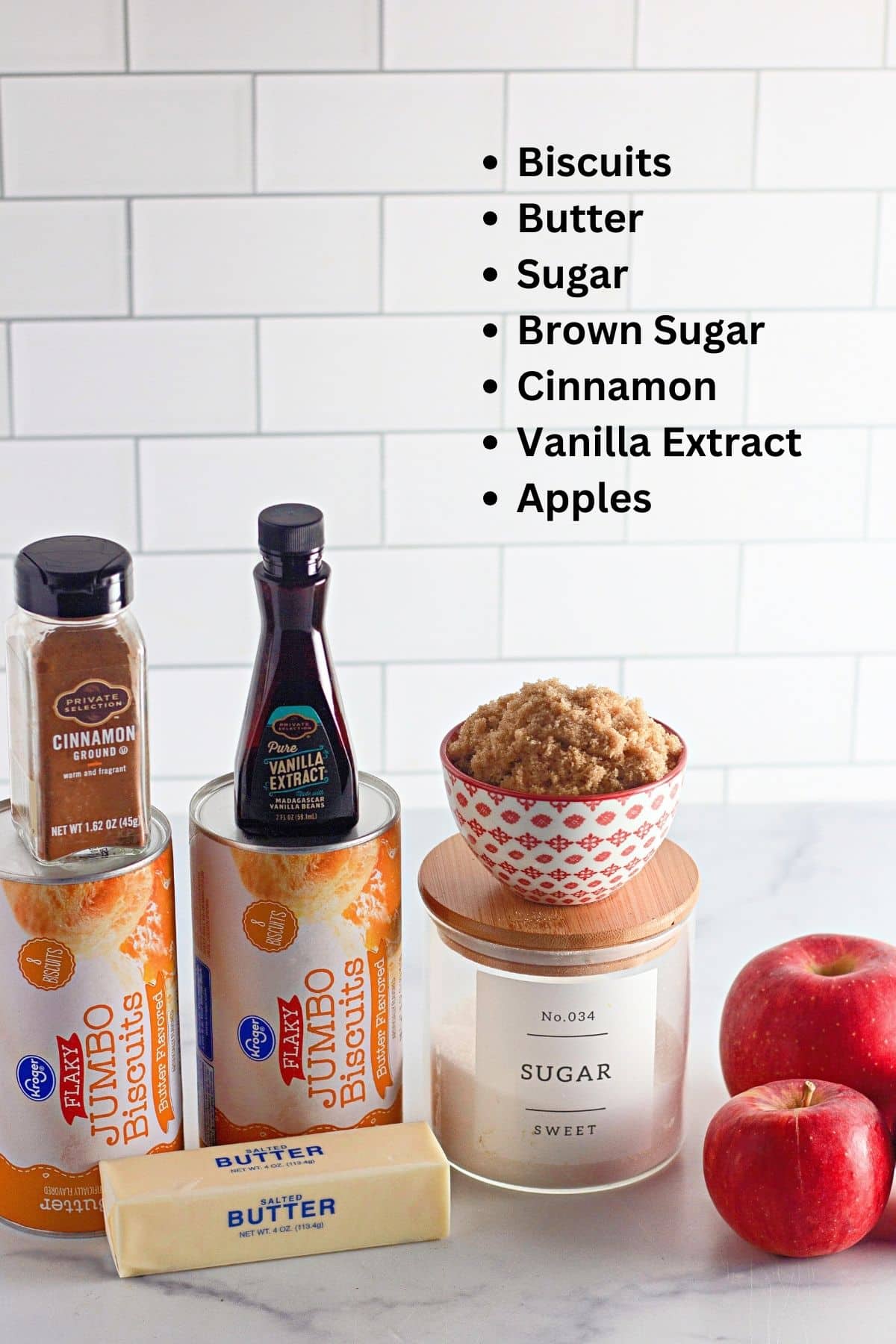 Ingredients for baking displayed on a counter, including biscuits, butter, brown sugar, cinnamon, vanilla extract, sugar, and apples.