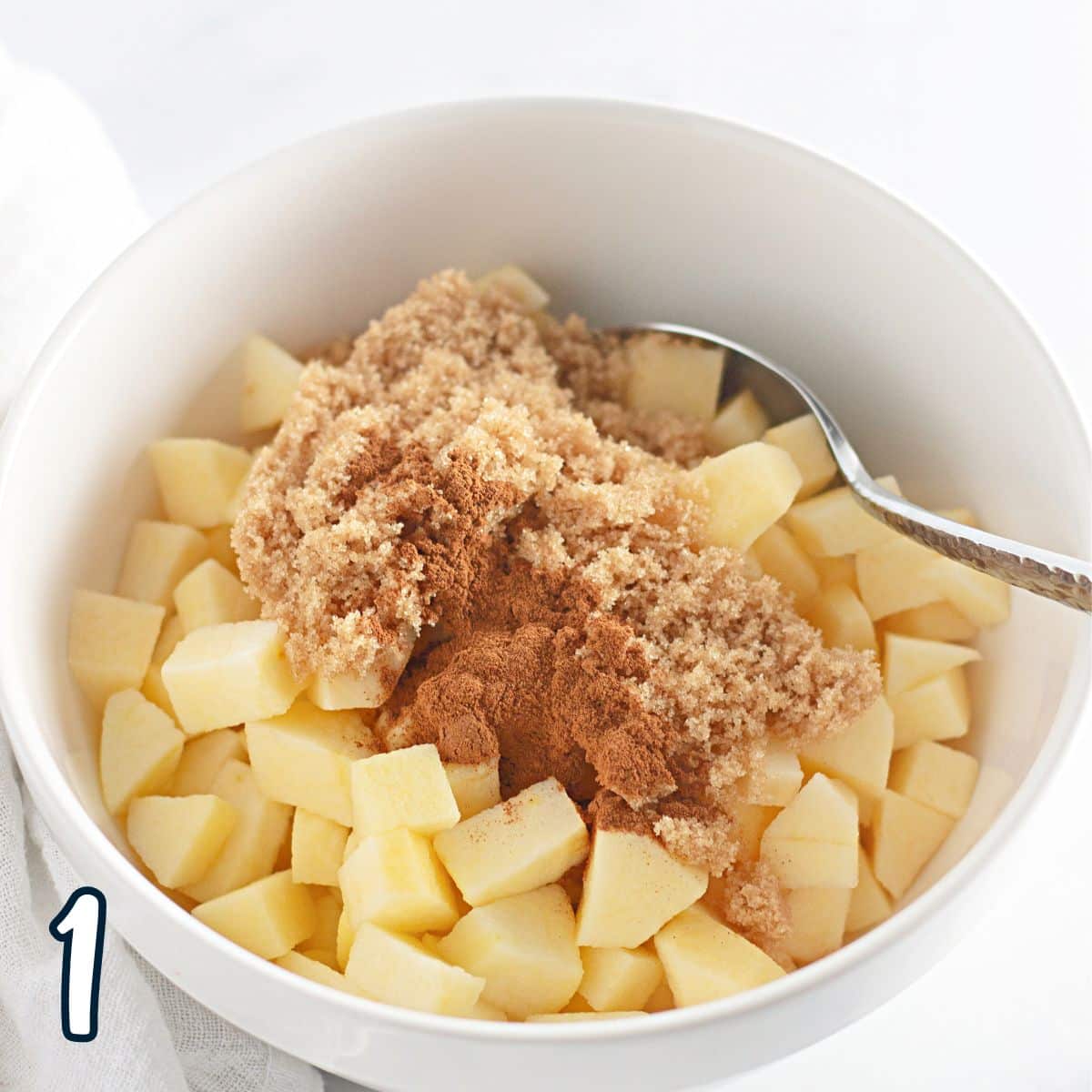 A bowl containing diced apple pieces sprinkled with brown sugar and cinnamon, with a spoon.