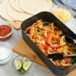 A dish of chicken fajitas with sliced bell peppers in a black pan, accompanied by tortillas, shredded cheese, salsa, and sour cream on a kitchen counter.