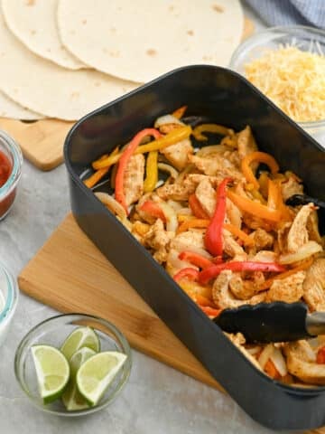 A dish of chicken fajitas with sliced bell peppers in a black pan, accompanied by tortillas, shredded cheese, salsa, and sour cream on a kitchen counter.