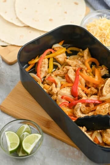 A dish of chicken fajitas with sliced bell peppers in a black pan, accompanied by tortillas, shredded cheese, salsa, and sour cream on a kitchen counter.