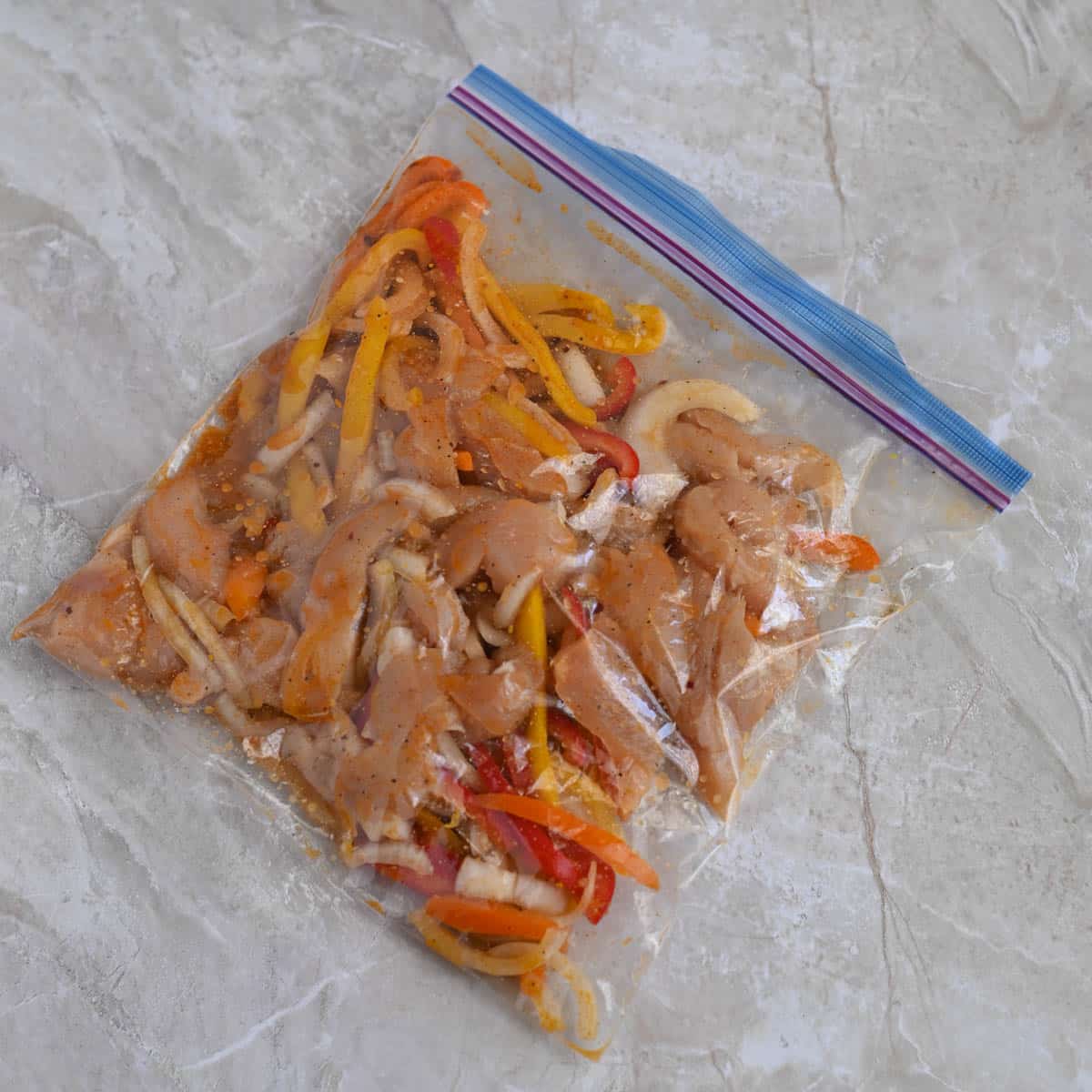 Chicken and sliced vegetables marinated in sauce inside a sealed plastic bag, placed on a textured surface.