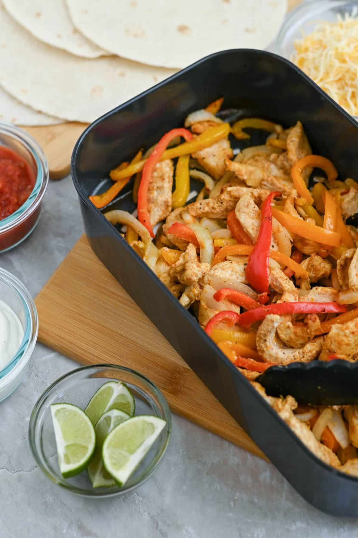 A dish of chicken fajitas with sliced bell peppers, served with sides of shredded cheese, salsa, sour cream, lime wedges, and tortillas.
