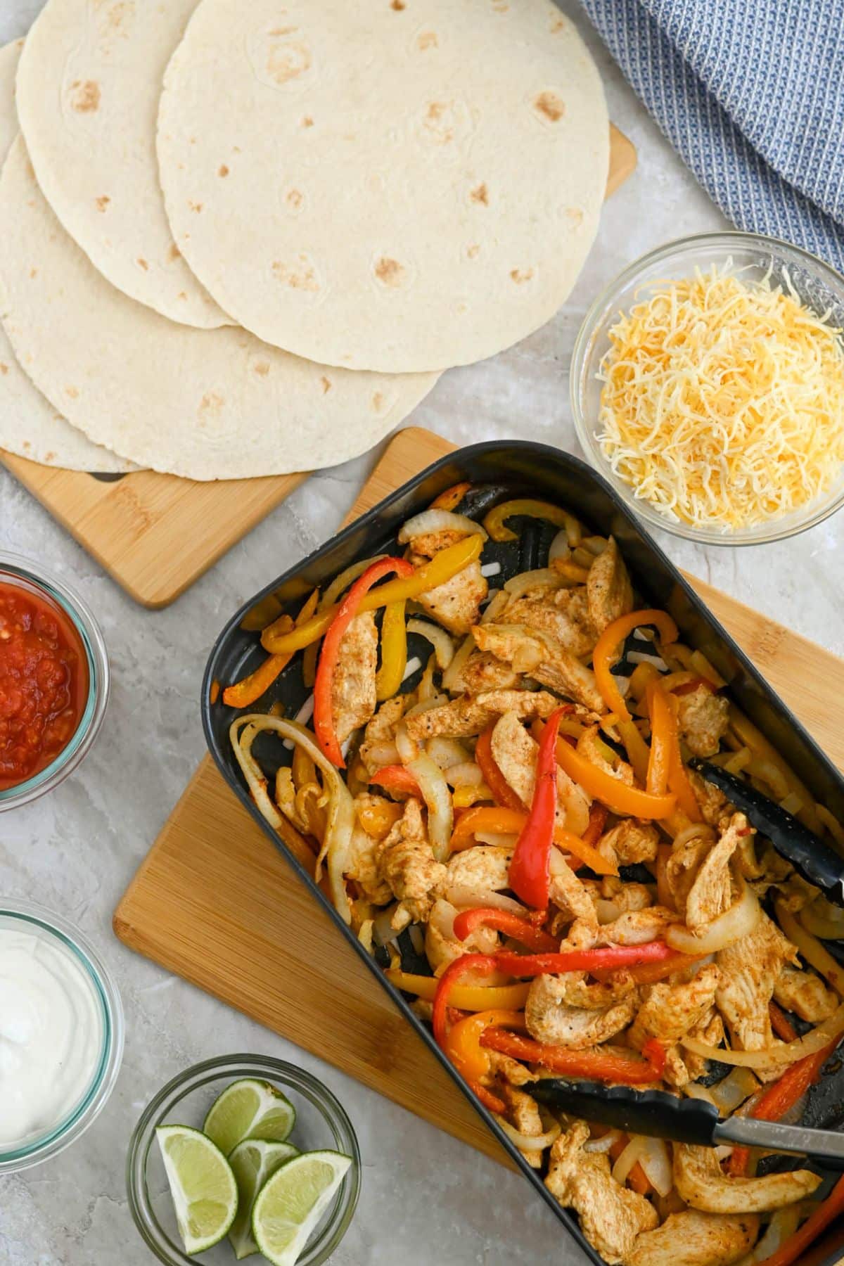 Top view of a chicken fajita meal with sliced bell peppers and onions in a skillet, surrounded by tortillas, cheese, salsa, sour cream, and lime wedges.