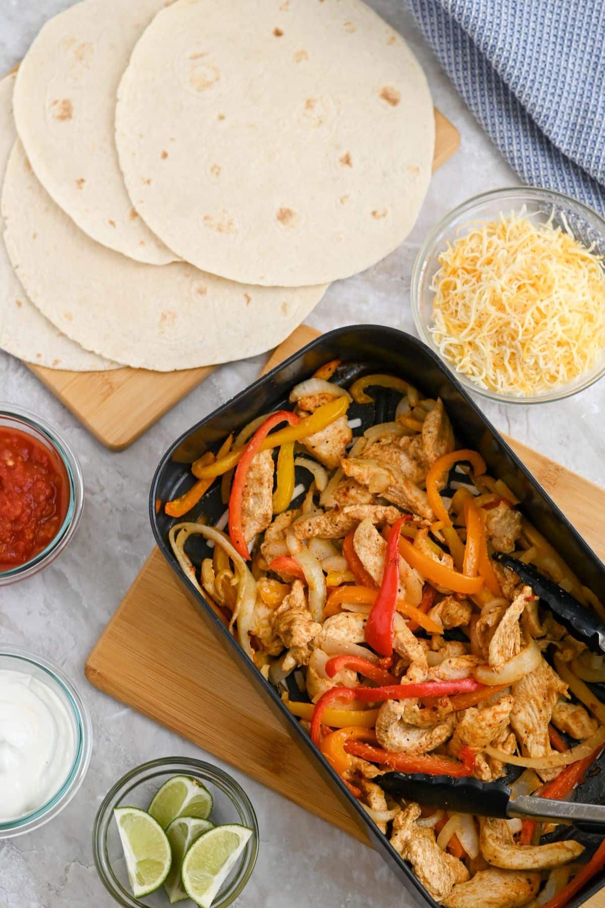 A top view of a chicken fajita mix with sliced bell peppers and onions in a black dish, surrounded by tortillas, shredded cheese, salsa, and lime on a marble countertop.