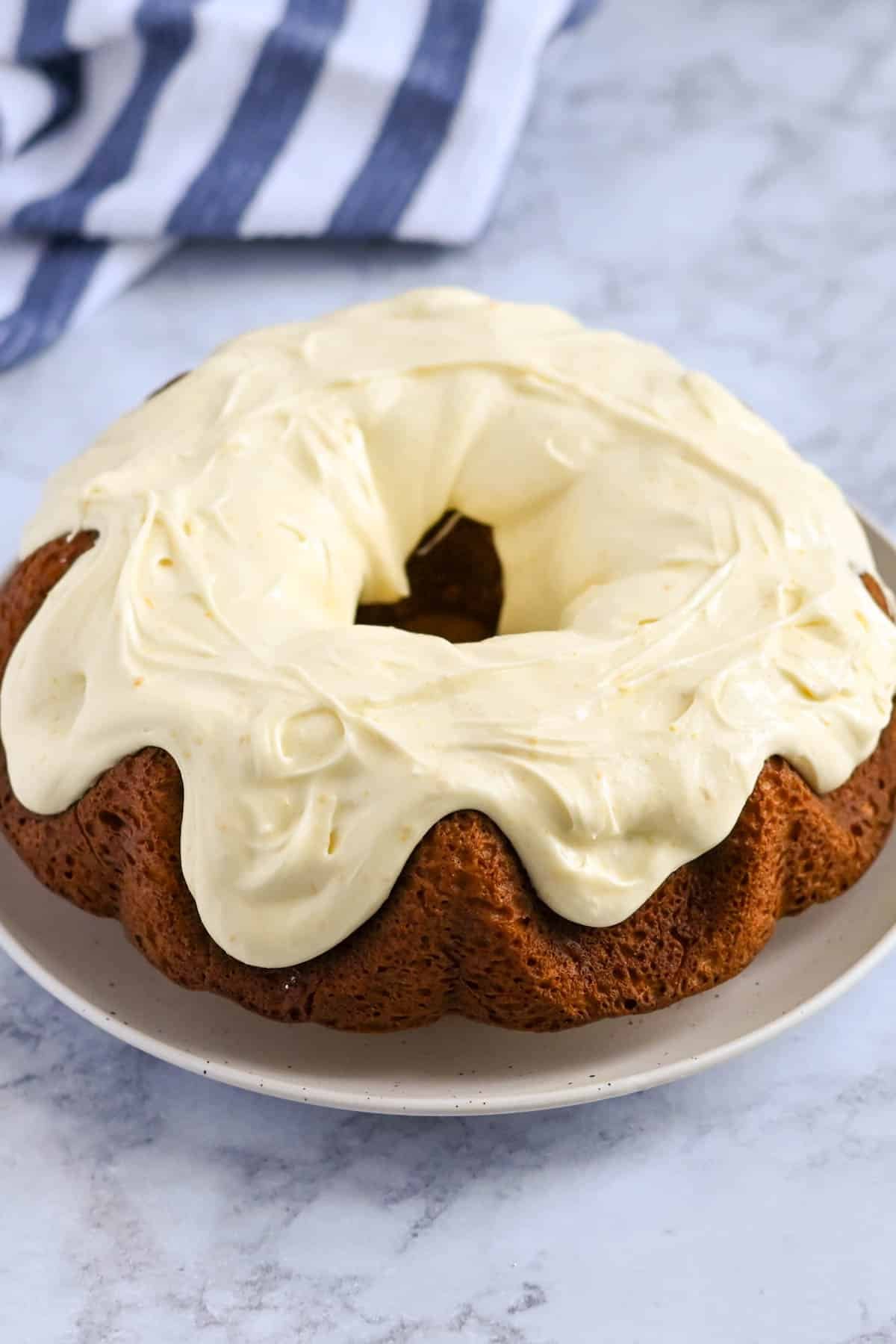 A Bundt cake with cream cheese frosting sits on a white plate on a marble surface, with a blue and white striped cloth in the background.