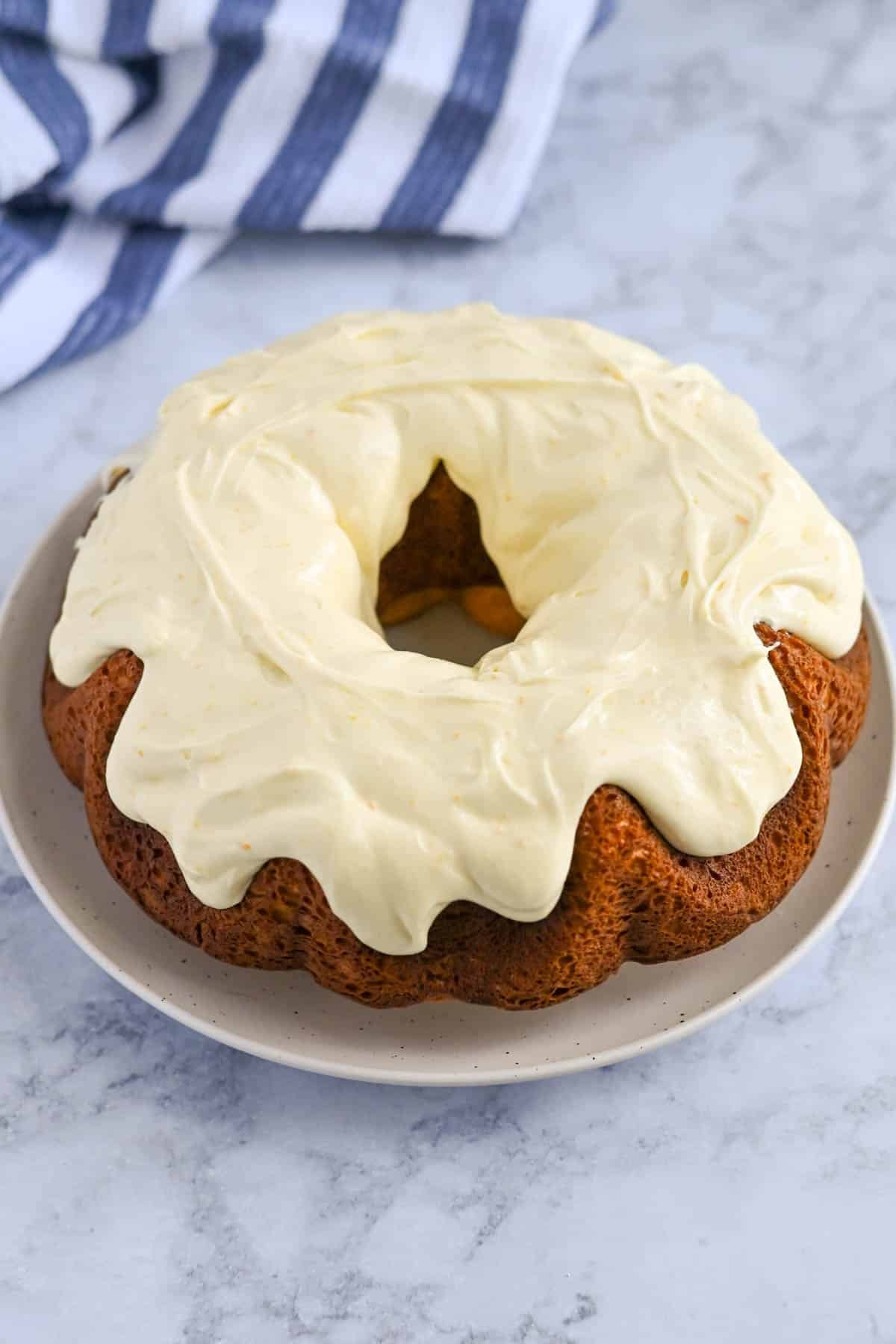 A Bundt cake with white frosting on top sits on a white plate, placed on a marble surface. A striped cloth is in the background.