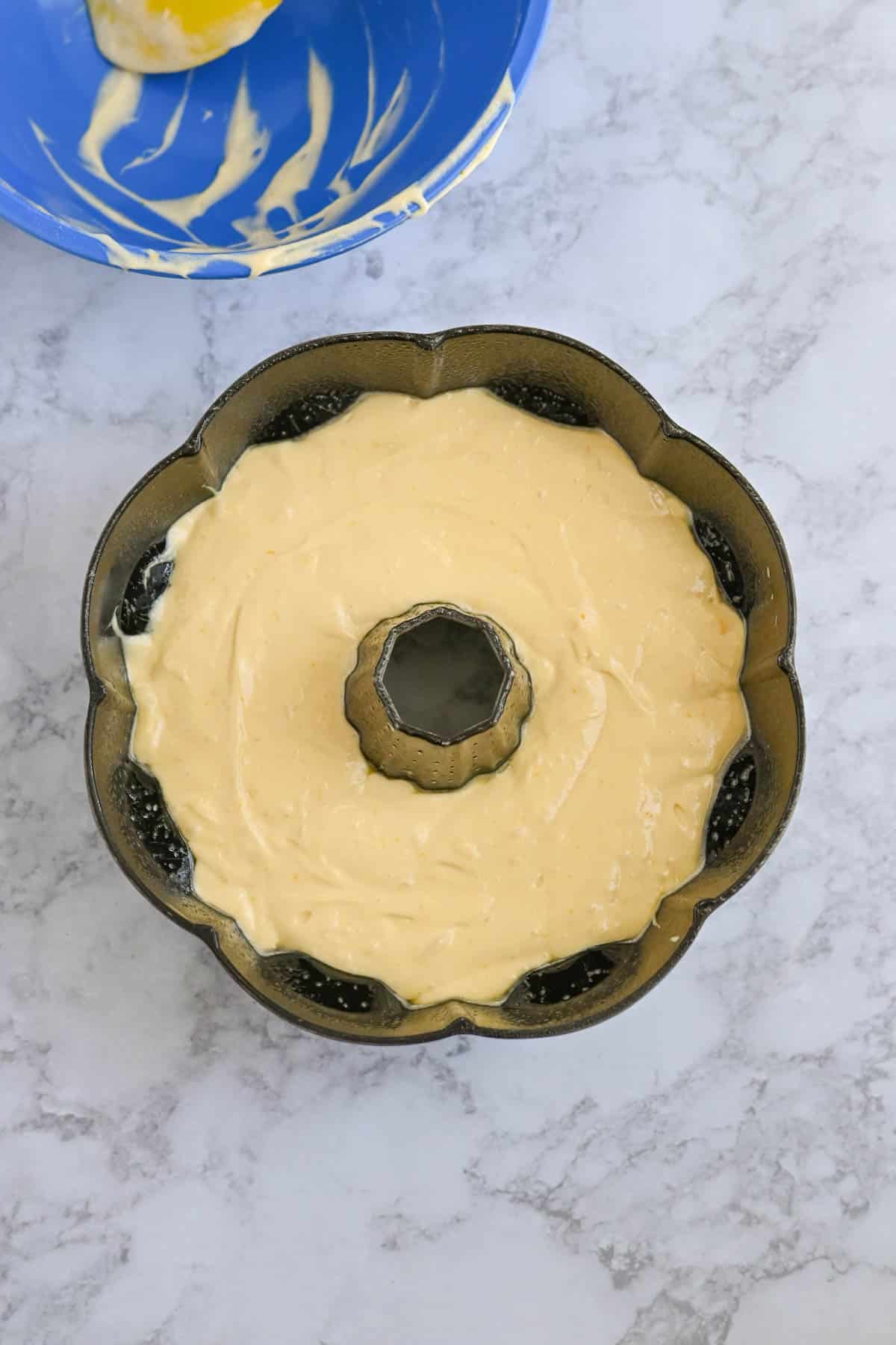 A bundt pan filled with cake batter is placed on a marble countertop. A partially empty blue bowl with remnants of batter is in the background.