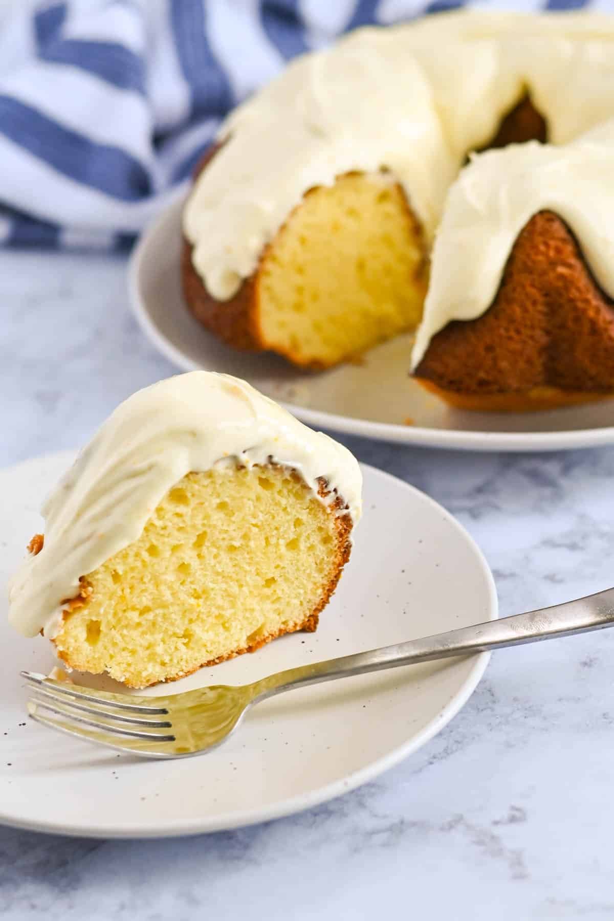 A slice of orange cake with cream cheese frosting rests on a white plate with a fork, while the rest of the frosted bundt cake sits on a second plate in the background.