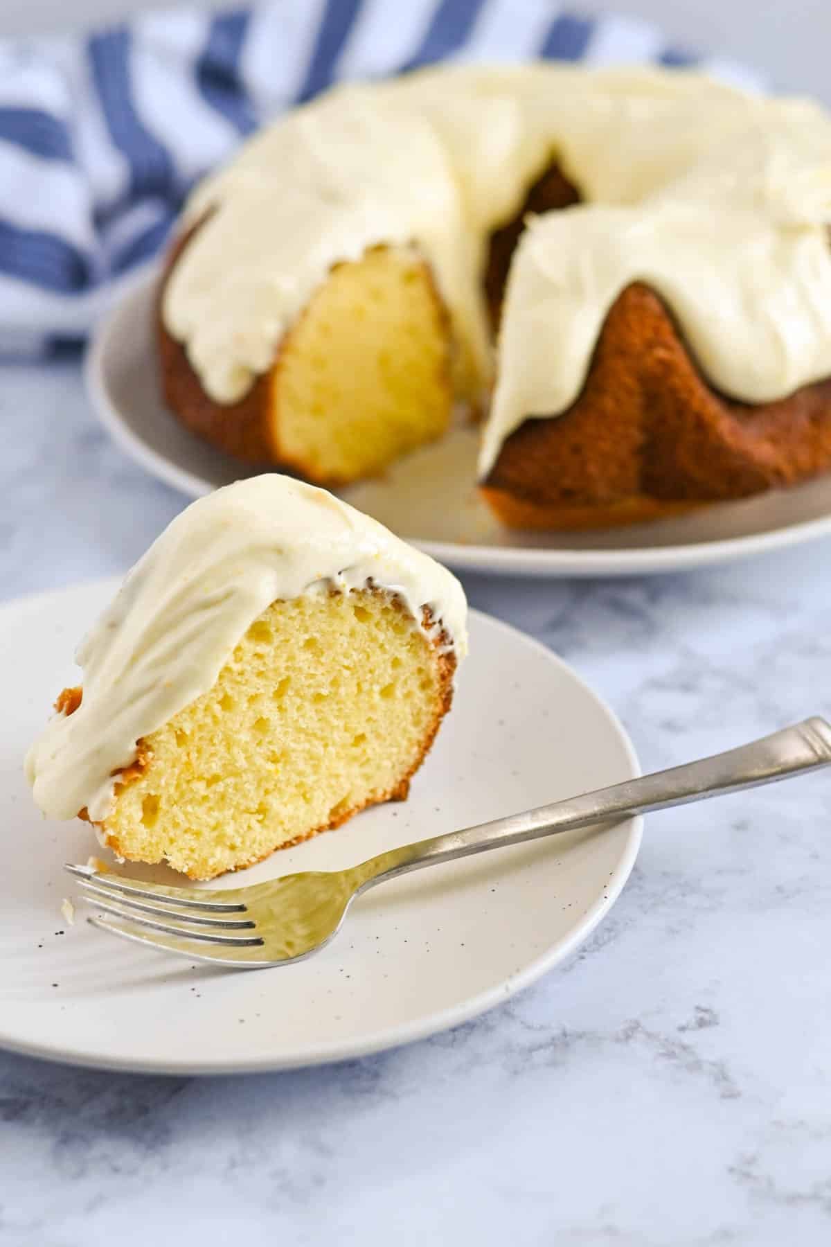 Slice of frosted bundt cake on a plate with a fork; whole bundt cake with a missing slice in the background.