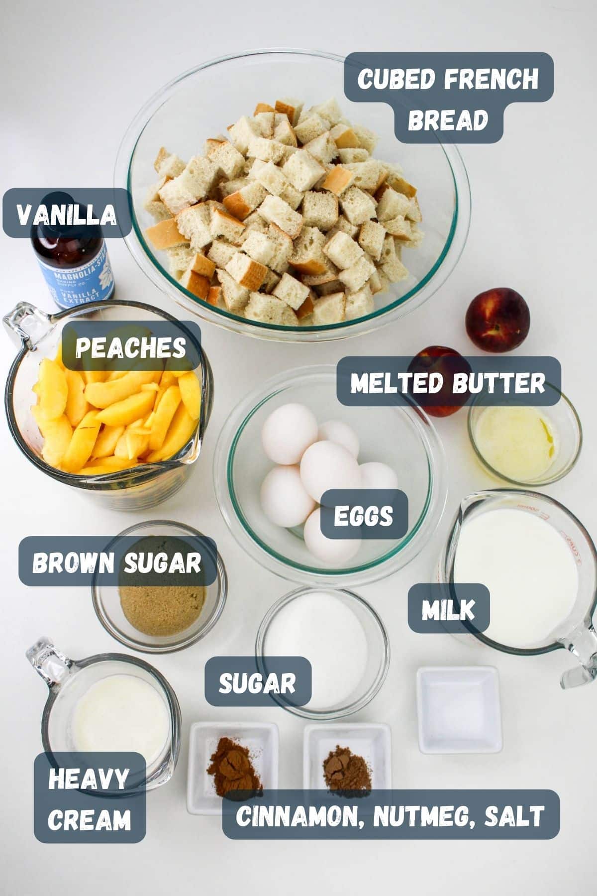 Ingredients for peach French Toast arranged on a counter, including cubed French bread, peaches, vanilla, melted butter, brown sugar, eggs, milk, heavy cream, sugar, and spices (cinnamon, nutmeg, salt).