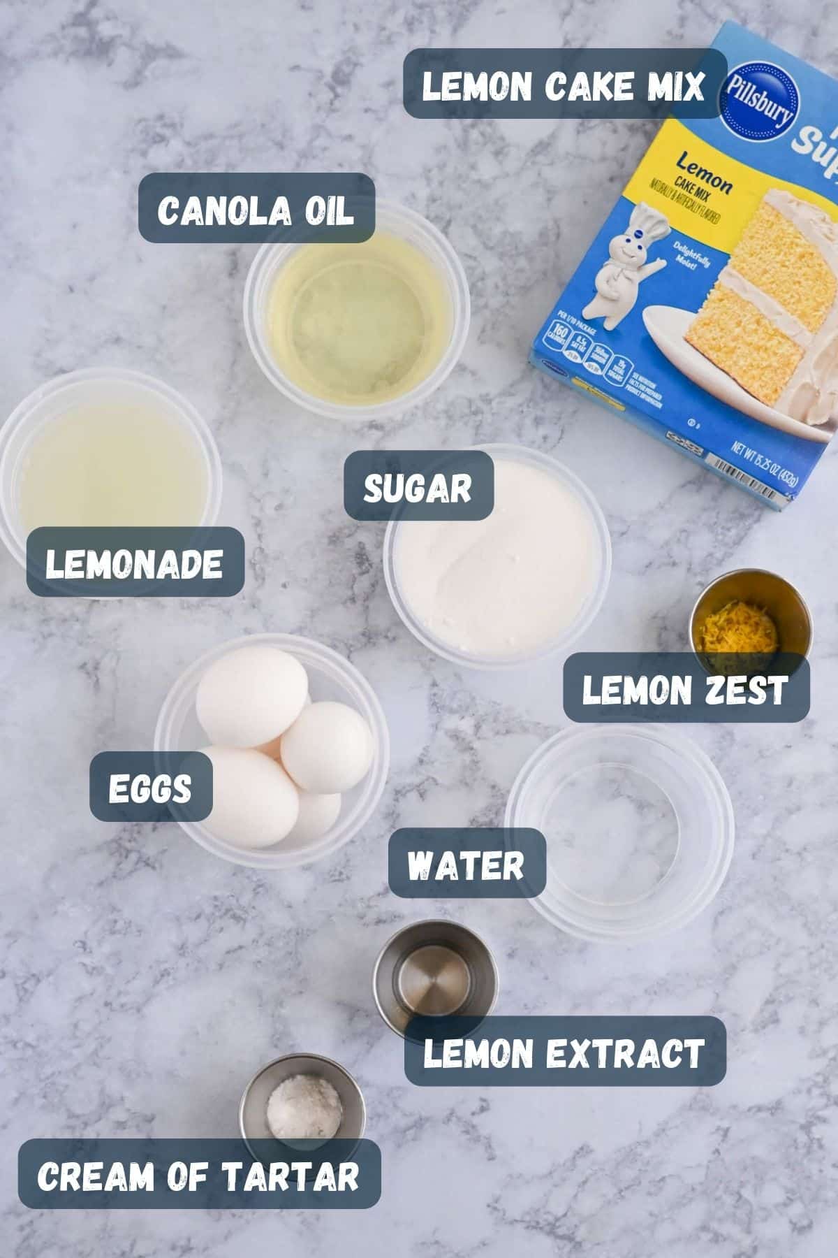 Ingredients for a lemon cake recipe include lemon cake mix, canola oil, lemonade, sugar, lemon zest, cream of tartar, lemon extract, eggs, and water, displayed on a marble surface.