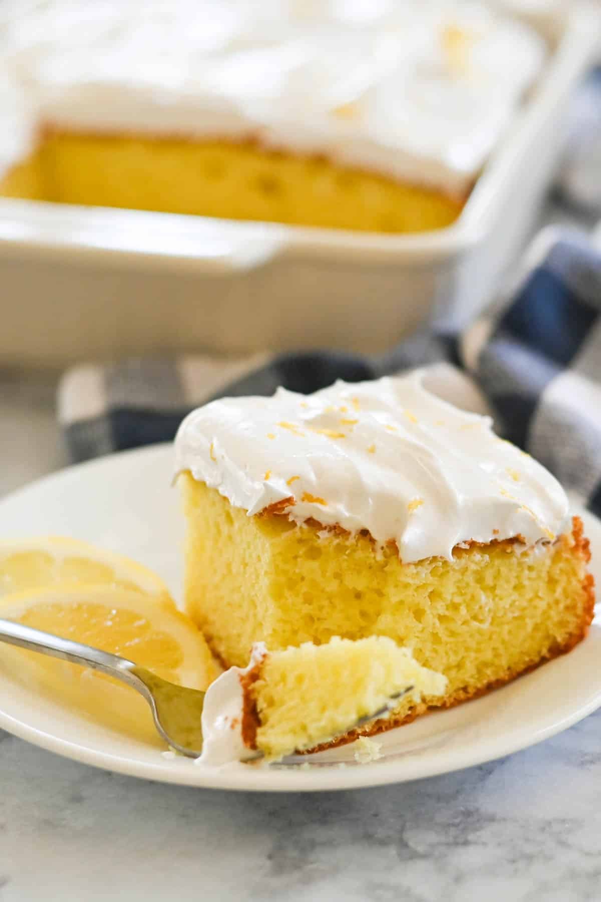 A slice of yellow cake with white frosting on a plate next to lemon slices, with a fork holding a piece of the cake. A pan with the remaining cake is in the background.