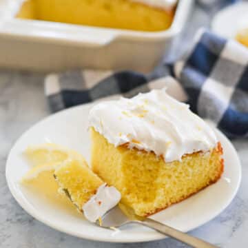 A square slice of frosted yellow cake is served on a white plate with a fork holding a piece. A blue and white checkered napkin is visible in the background.