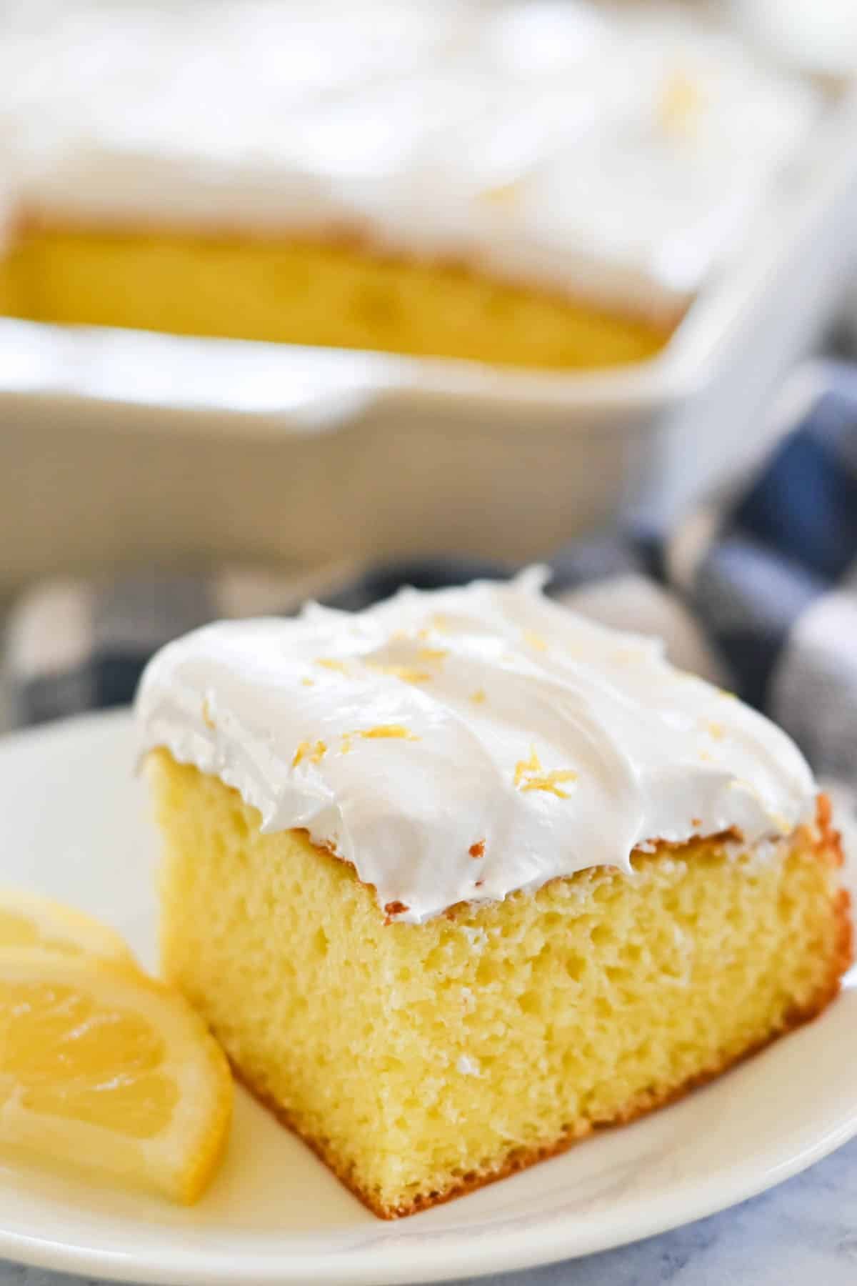 A slice of frosted yellow cake with a lemon garnish on a white plate. A larger cake in a white baking dish is in the background.