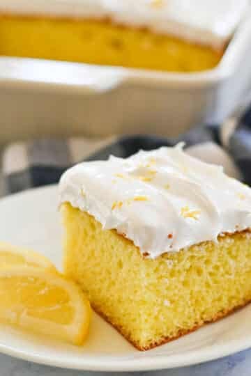 A slice of lemon cake topped with white frosting is served on a white plate with two lemon wedges, with a larger portion of the cake in the background.