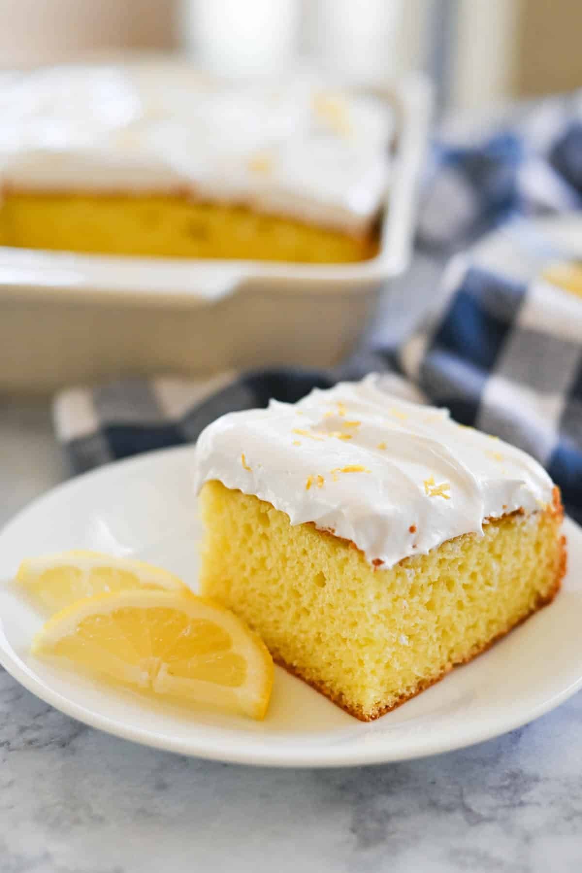 A slice of lemon cake with marshmallow frosting on a white plate, garnished with lemon slices. Blurred background shows the rest of the cake in a baking dish.