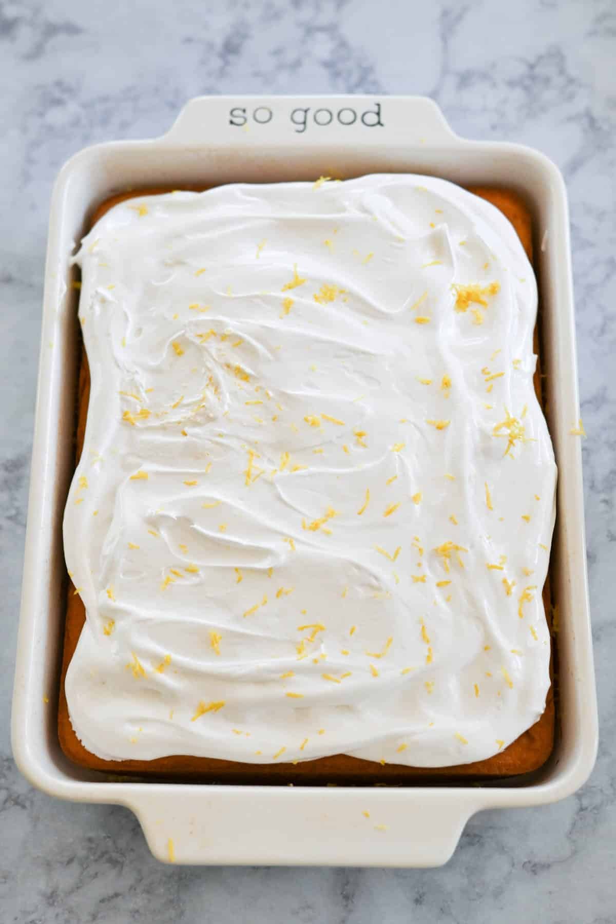 A rectangular cake with marshmallow frosting and lemon zest is placed in a white ceramic baking dish, which has "so good" inscribed on one of its handles. The dish rests on a marble surface.