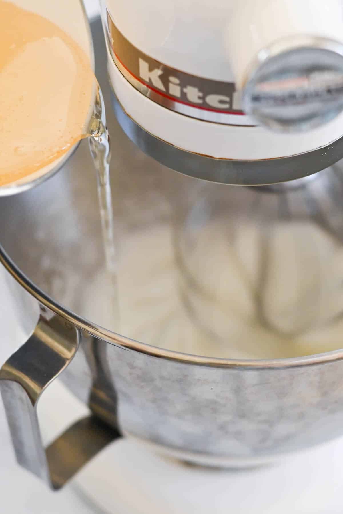 A liquid is being poured into a stand mixer with a stainless steel bowl, while the mixer attachment is visible inside the bowl.