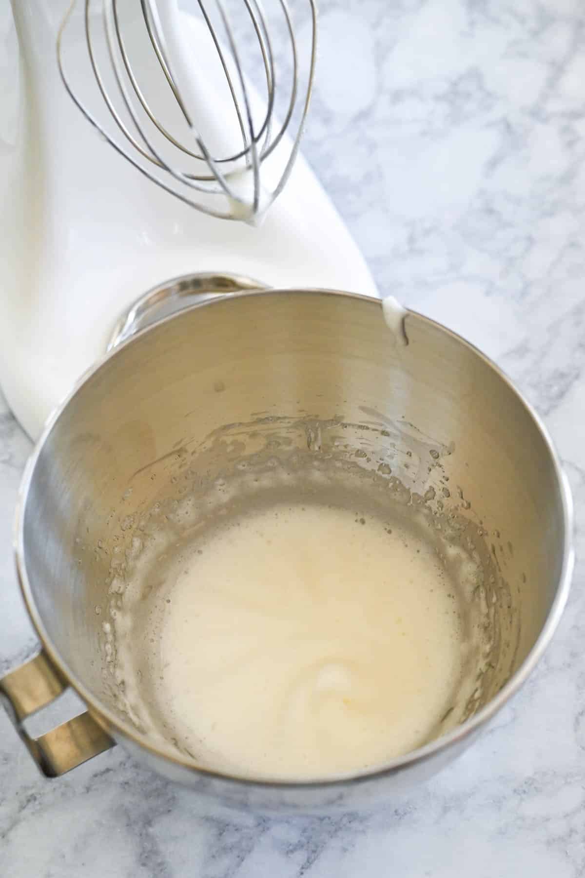 A stand mixer with a whisk attachment over a metal bowl containing partially whipped egg whites on a marble countertop.