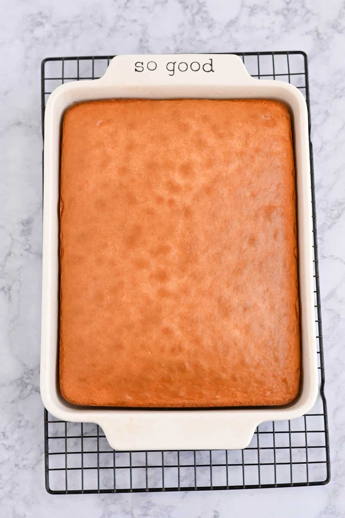 A rectangular cake in a white baking dish with the words "so good" written on the edge sits on a cooling rack.