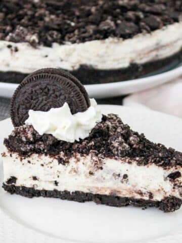 A slice of cookies and cream cheesecake topped with crushed cookies, whipped cream, and a whole cookie on a white plate.
