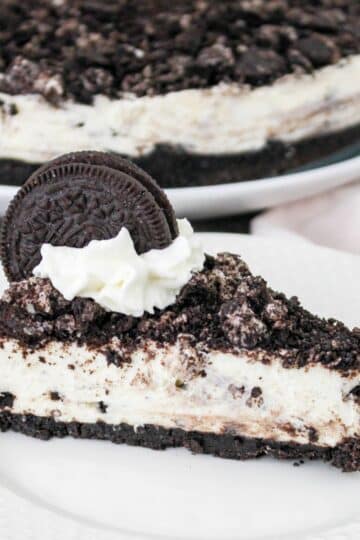 A slice of cookies and cream cheesecake topped with crushed cookies, whipped cream, and a whole cookie on a white plate.
