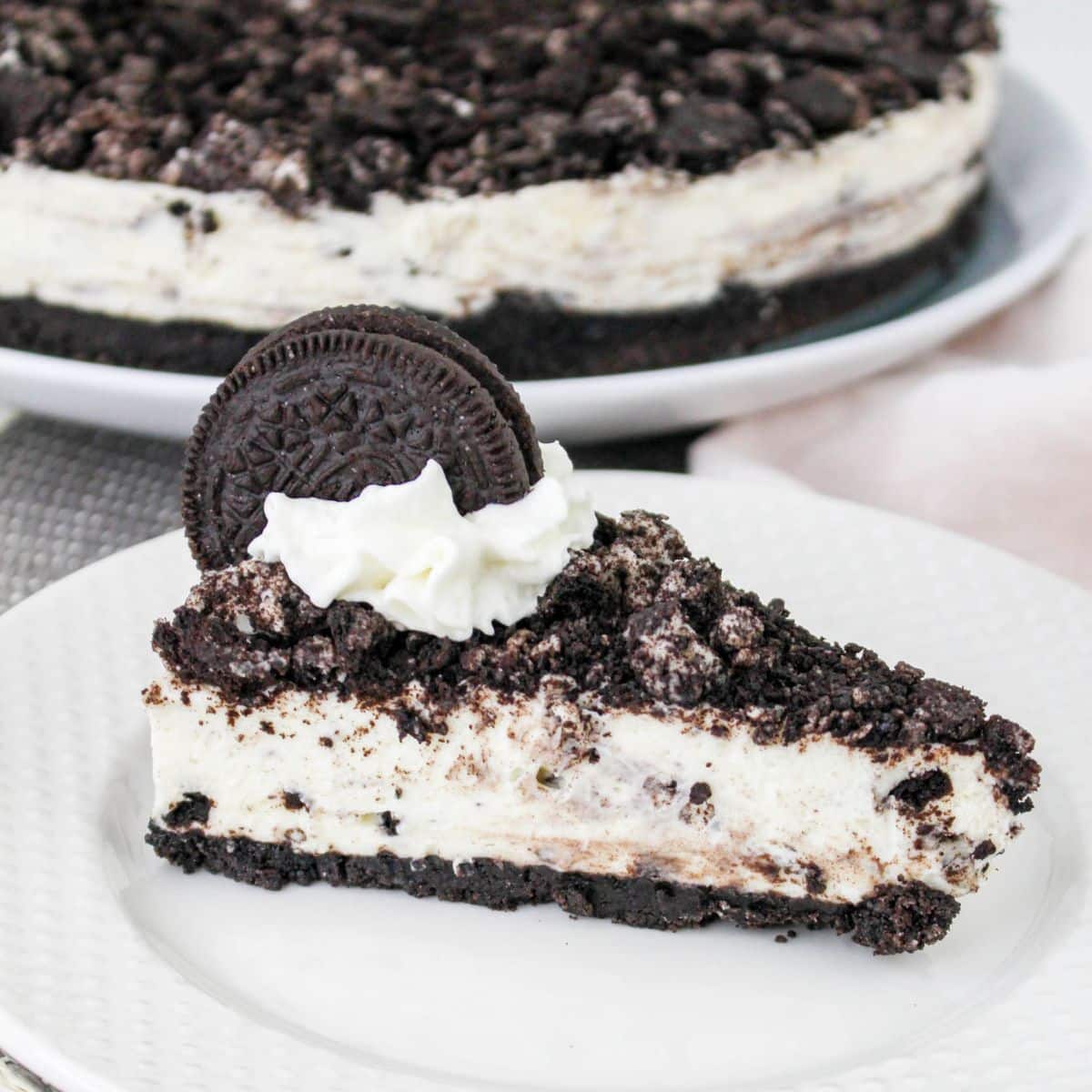 A slice of cookies and cream cheesecake topped with crushed cookies, whipped cream, and a whole cookie on a white plate.