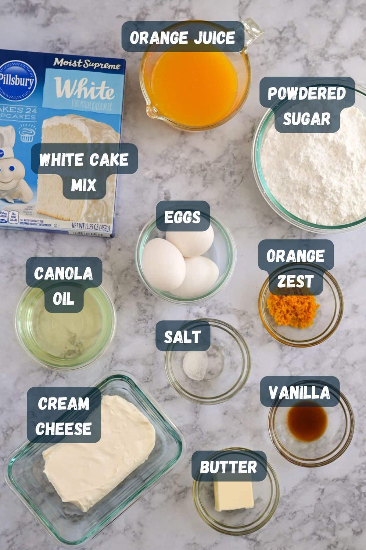A variety of baking ingredients are laid out on a marble surface. Labels identify them as white cake mix, powdered sugar, eggs, orange juice, orange zest, canola oil, cream cheese, butter, salt, and vanilla.
