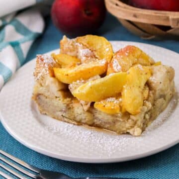 A square slice of peach baked French Toast with powdered sugar, is served on a white plate. There are peaches and a basket in the background.