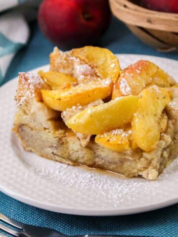 A square slice of peach baked French Toast with powdered sugar, is served on a white plate. There are peaches and a basket in the background.