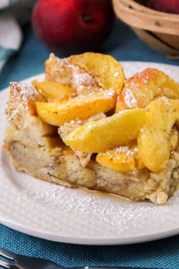 A square slice of peach baked French Toast with powdered sugar, is served on a white plate. There are peaches and a basket in the background.