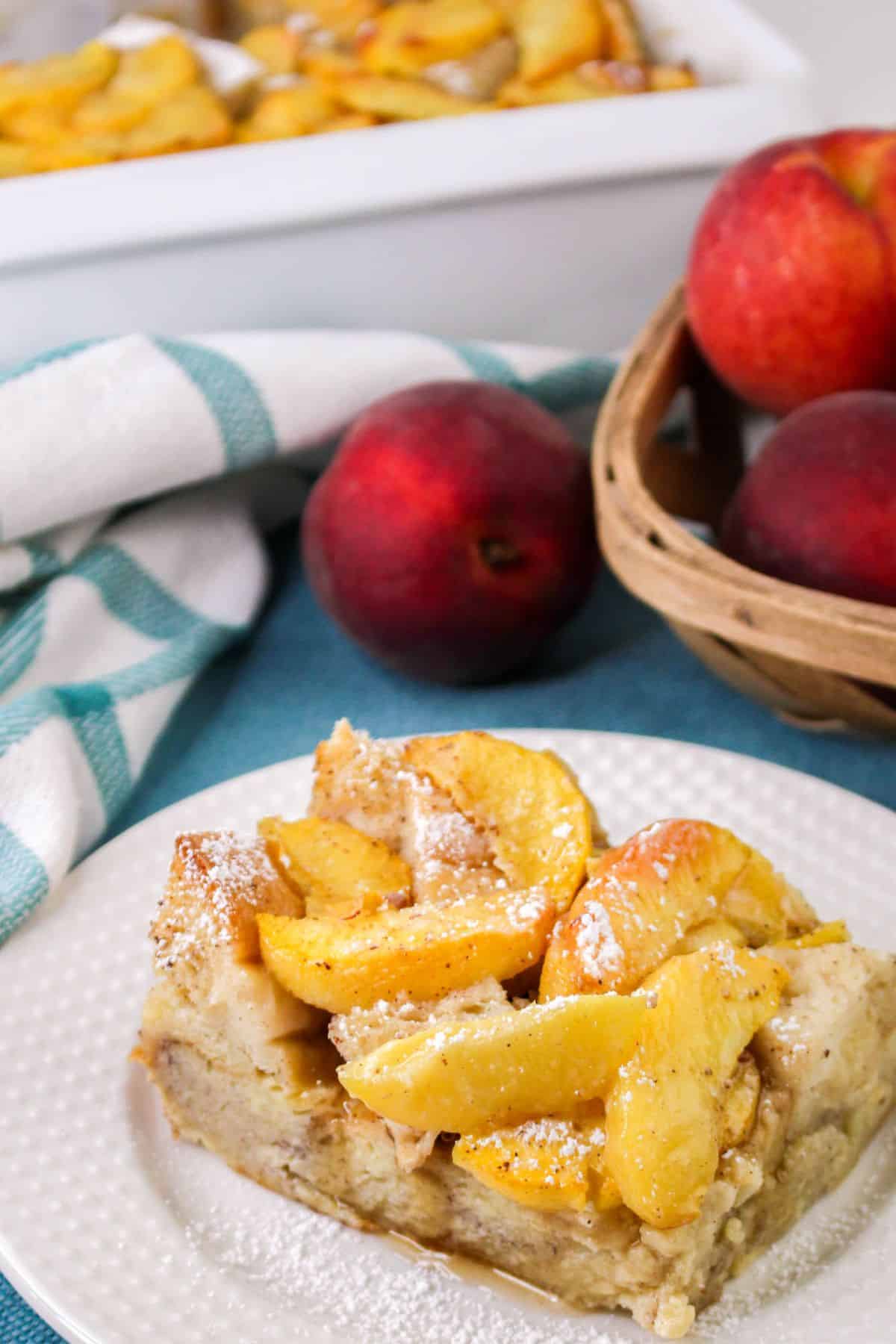 A plate with a piece of peach French Toast dusted with powdered sugar. In the background, there's a basket of whole peaches and a baking dish with more of the dessert.