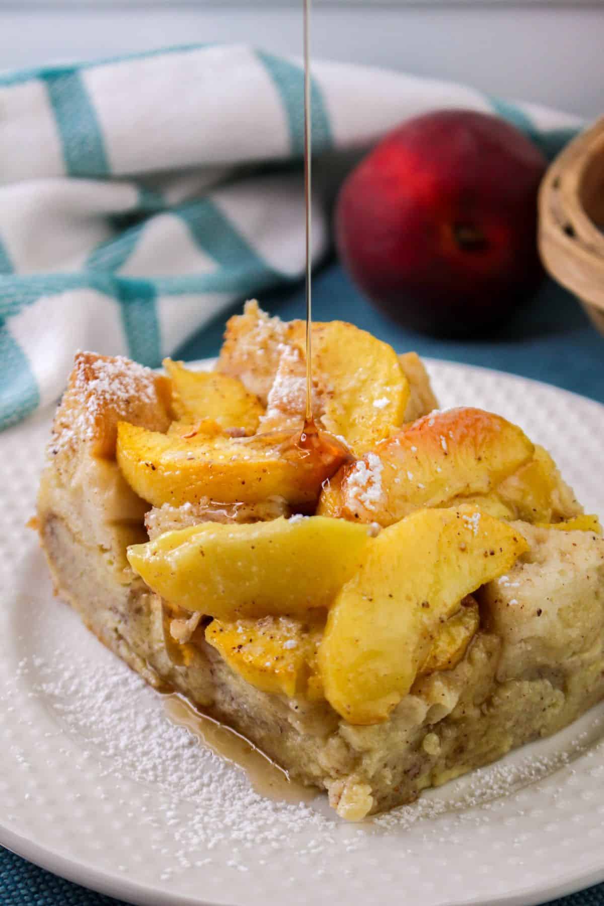 A slice of bread pudding topped with peach slices and powdered sugar is drizzled with syrup. A peach and a cloth are in the background.