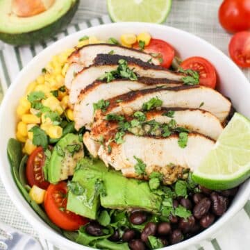 A bowl of salad with grilled chicken, black beans, corn, avocado, cherry tomatoes, and lime, garnished with cilantro.