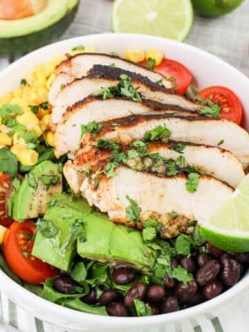 A bowl of salad with grilled chicken, avocado, tomatoes, black beans, corn, greens, and lime slices.