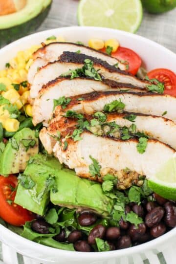 A bowl of salad with grilled chicken, avocado, tomatoes, black beans, corn, greens, and lime slices.