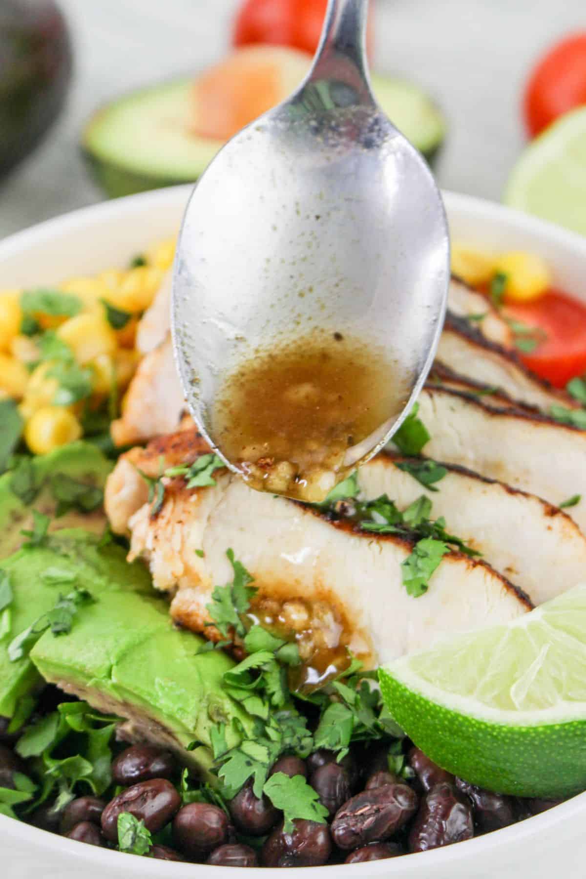 Grilled chicken salad in a bowl with black beans, avocado, corn, and lime being drizzled with dressing from a spoon.