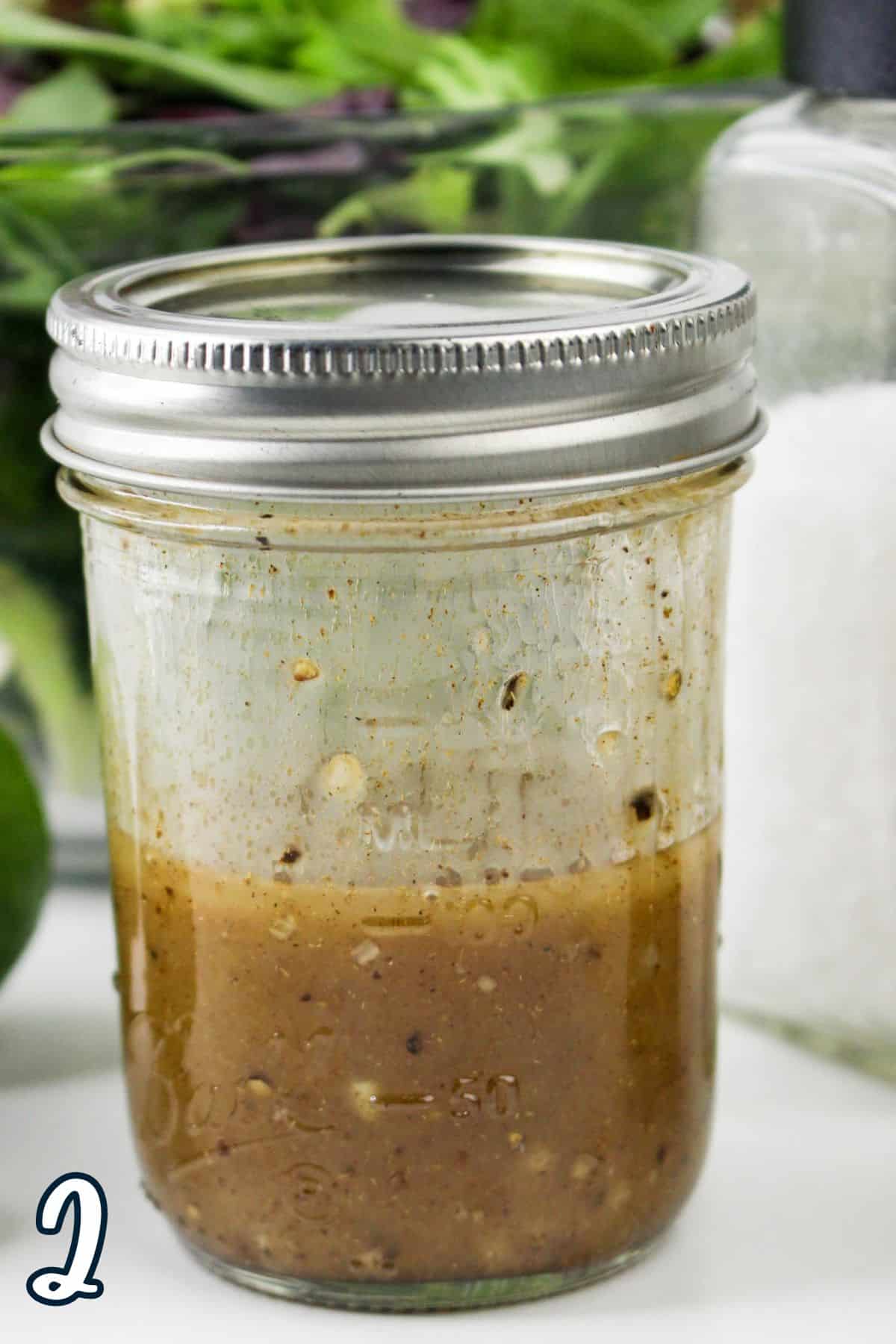 A jar of homemade salad dressing with visible spices sits next to salt, a jar, and greens in the background.