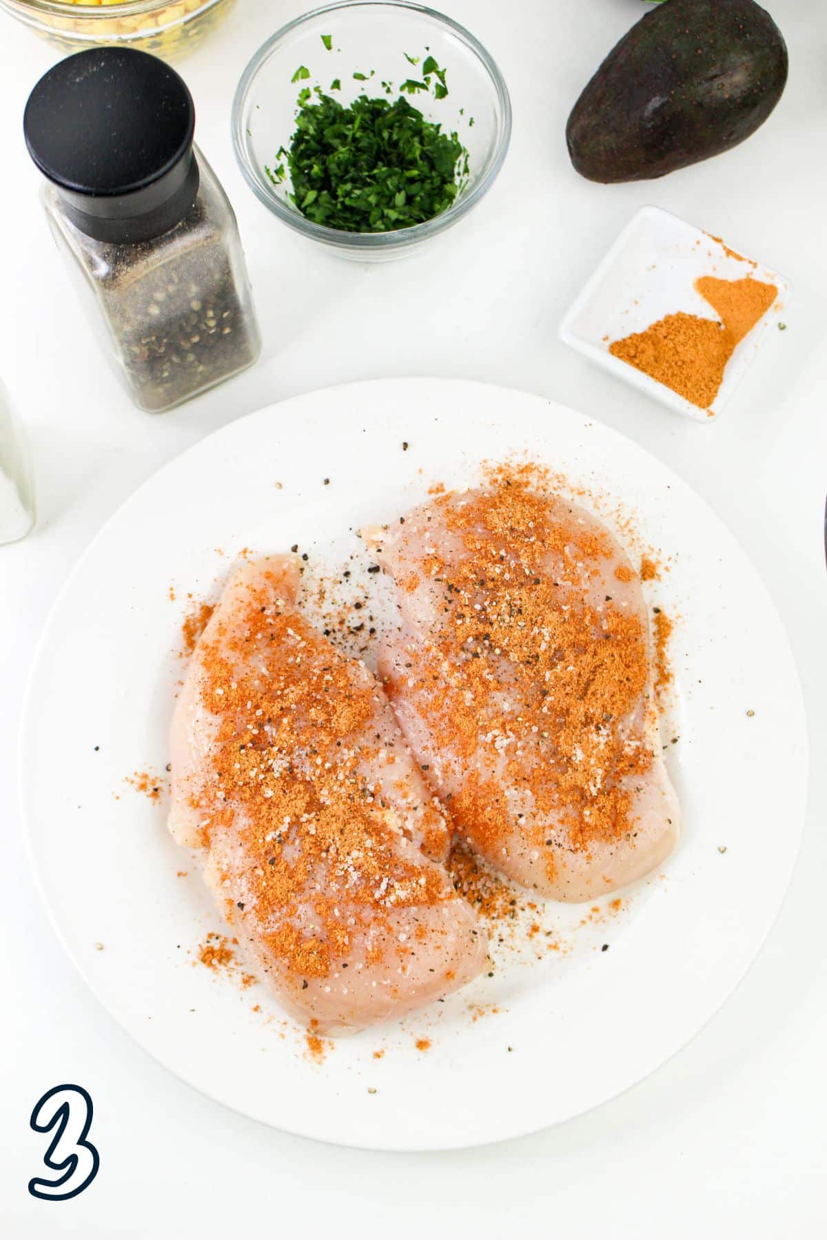 Raw chicken breasts with seasoning on a plate, surrounded by various spices and herbs on a white surface. The number "3" is displayed.