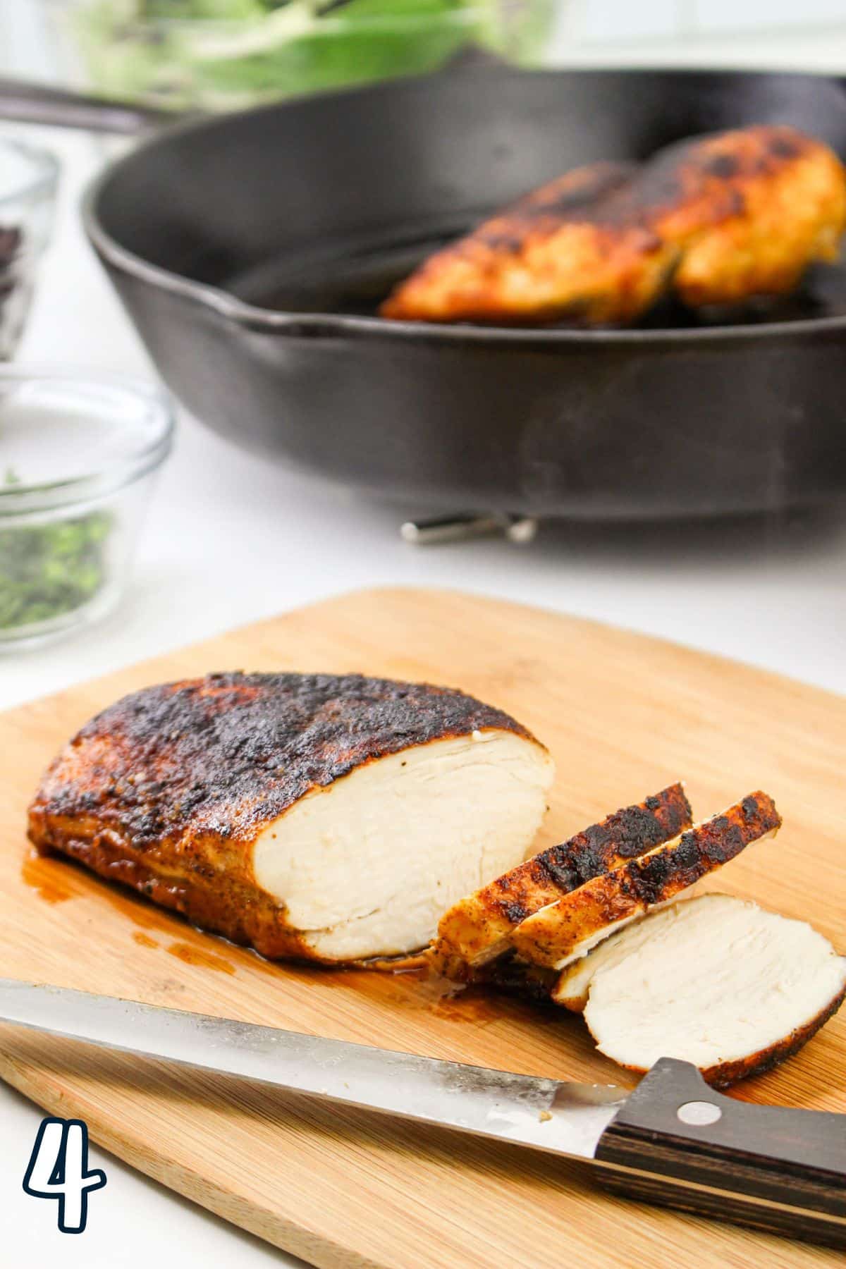 Sliced, cooked chicken breast on a cutting board with a knife, with a skillet containing another chicken breast in the background.